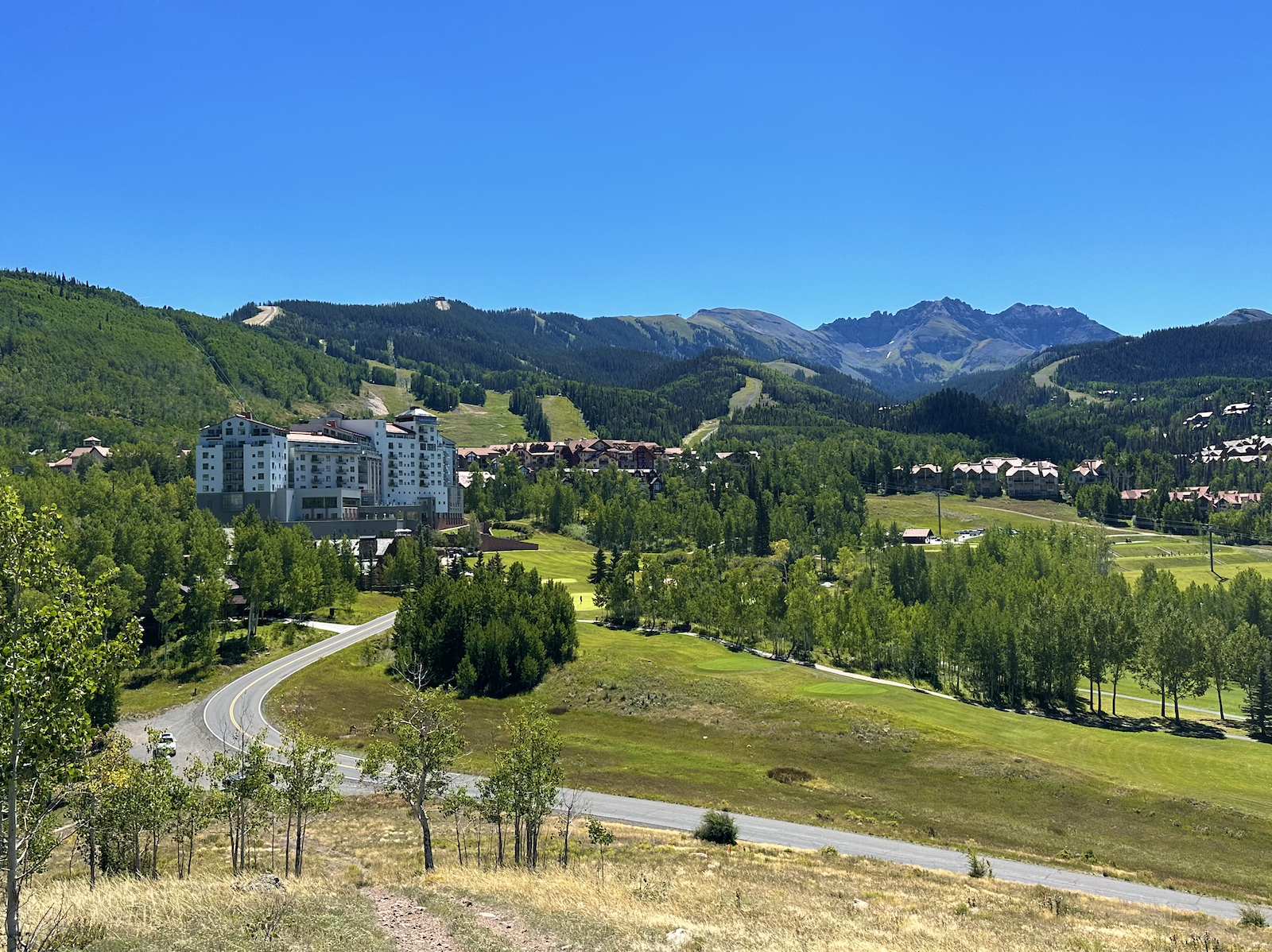 Mountain Village, Colorado in summer — alpine residences set among green forested slopes beneath the San Sophia range under a clear blue sky